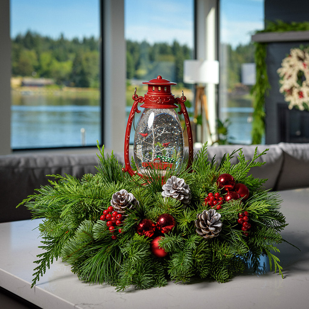 An arrangement of noble fir, incense cedar, and white pine with frosted pinecones, red berry clusters, red ball clusters, and a wintery scene in a red globe lantern sitting on a counter.