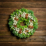 Holiday wreath made of noble fir, incense cedar, white pine, and bay leaf with faux white berries and leaves, and Australian pine cones hanging on a wooden wall