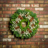 Holiday wreath made of noble fir, incense cedar, white pine, and bay leaf with faux white berries and leaves, and Australian pine cones hanging on a brick wall