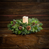 An arrangement of noble fir, cedar, white pine, salal, bay leaves, pinecones, and an ivory LED candle sitting on a wooden shelf with a dark wood background.