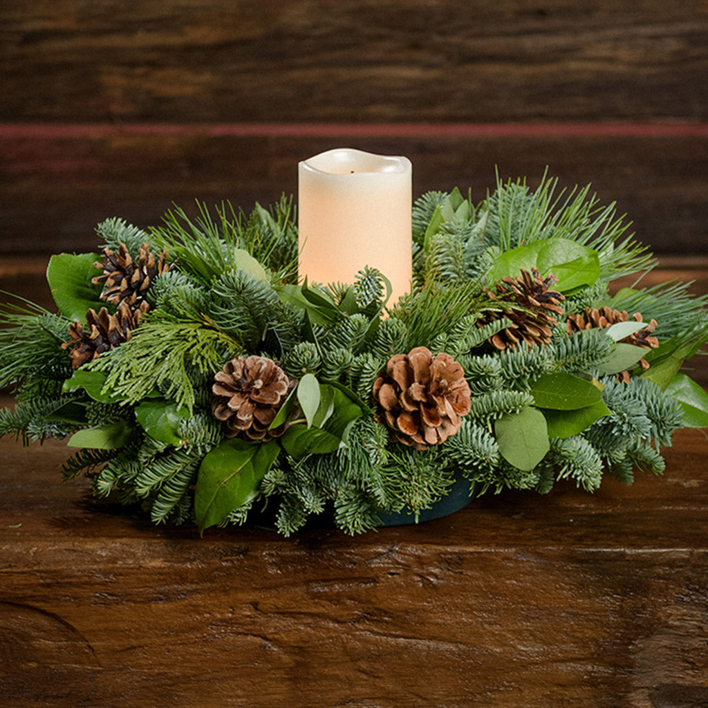 An arrangement of noble fir, cedar, white pine, salal, bay leaves, pinecones, and an ivory LED candle sitting on a wooden shelf with a dark wood background.