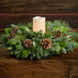 An arrangement of noble fir, cedar, white pine, salal, bay leaves, pinecones, and an ivory LED candle sitting on a wooden shelf with a dark wood background.