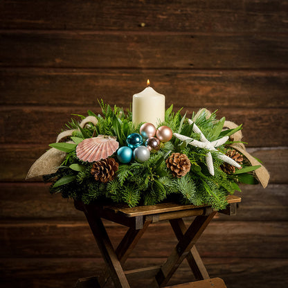 An arrangement made of noble fir, white pine, incense cedar, and bay leaves, with a seashell, seastar, aqua ball clusters, champagne ball clusters, Austrian pinecones, an ivory pillar candle, and tan linen ribbons sitting on a table with a dark wooden background.