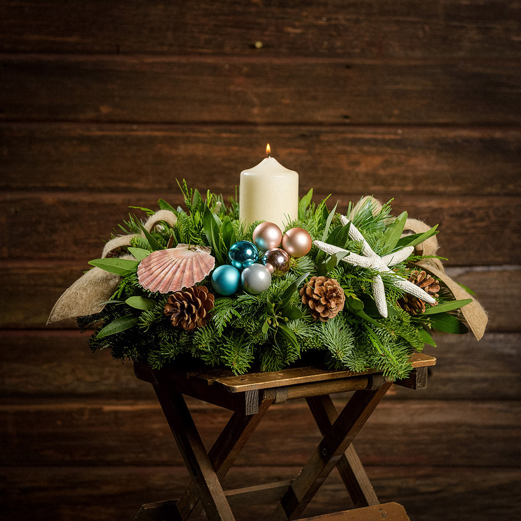 An arrangement made of noble fir, white pine, incense cedar, and bay leaves, with a seashell, seastar, aqua ball clusters, champagne ball clusters, Austrian pinecones, an ivory pillar candle, and tan linen ribbons sitting on a table with a dark wooden background.