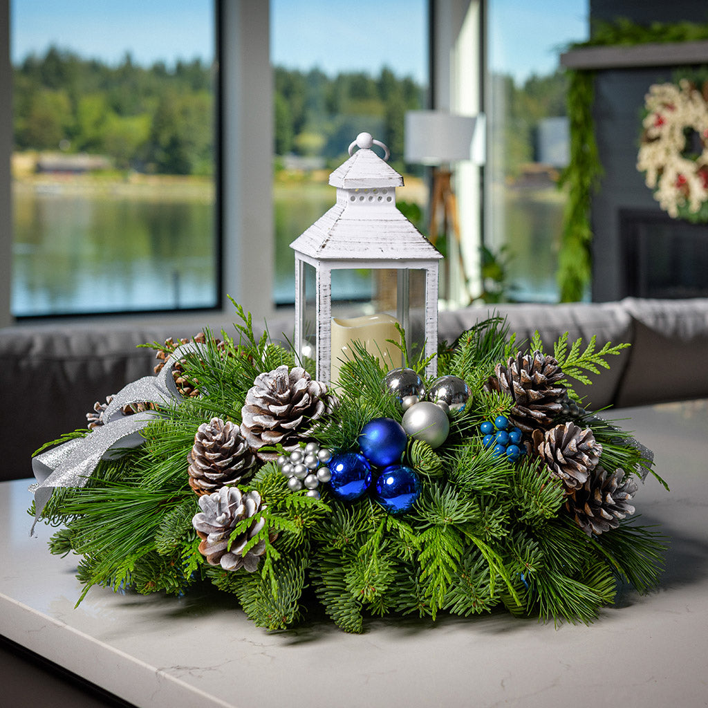 An arrangement made of noble fir, incense cedar, and white pine with ponderosa pinecones, Austrian pinecones, blue and silver ball ornament clusters, blue and silver berry clusters, silver glitter bow tucks, and white LED lantern sitting on a counter.