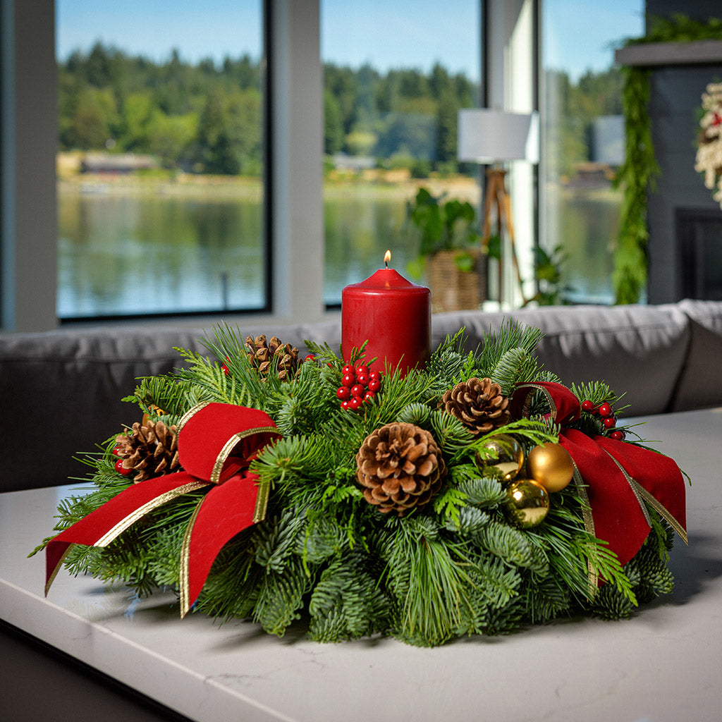 Centerpiece with pine cones, gold balls, red berry clusters,gold trimmed red bow tucks and a red pillar candle sitting on a counter.