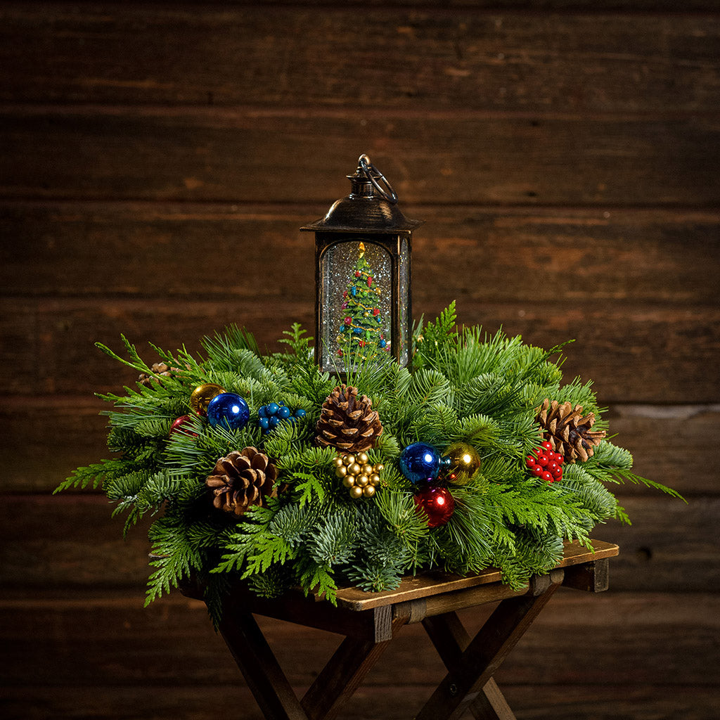 An arrangement made of noble fir, incense cedar, and white pine with Austrian pinecones, red, blue, and gold berry and ball clusters, and a musical Christmas tree spinning in a lantern globe sitting on a wood table with a dark wooden background.