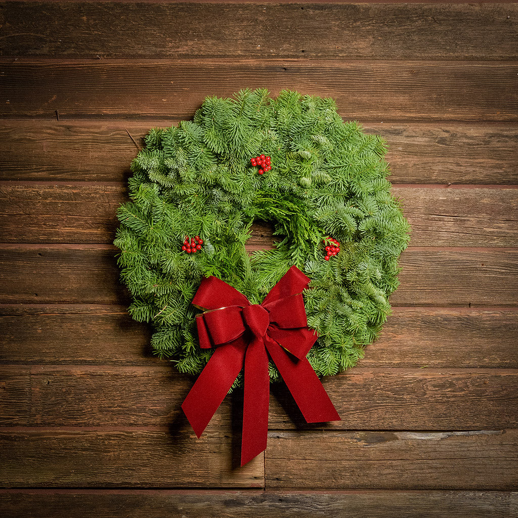 22" Christmas wreath of fir and cedar with three red berry clusters and a gold-backed red velveteen bow hanging on a white wood background.