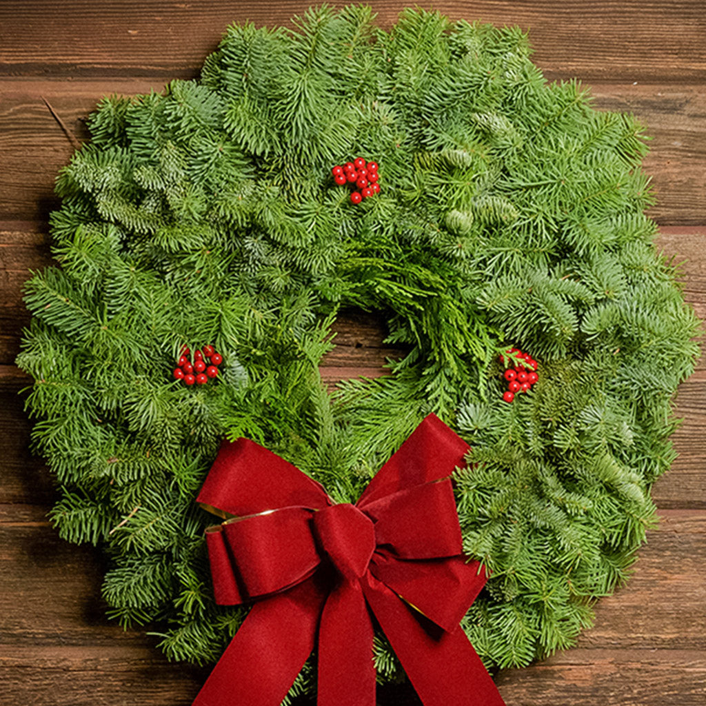 22" Christmas wreath of fir and cedar with three red berry clusters and a gold-backed red velveteen bow hanging on a dark wood background.