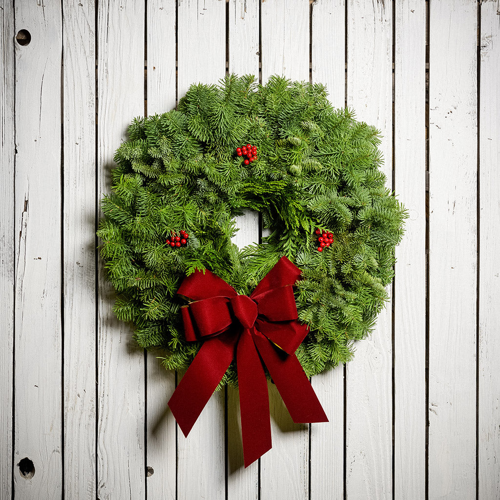 22" Christmas wreath of fir and cedar with three red berry clusters and a gold-backed red velveteen bow hanging on a white wood background.