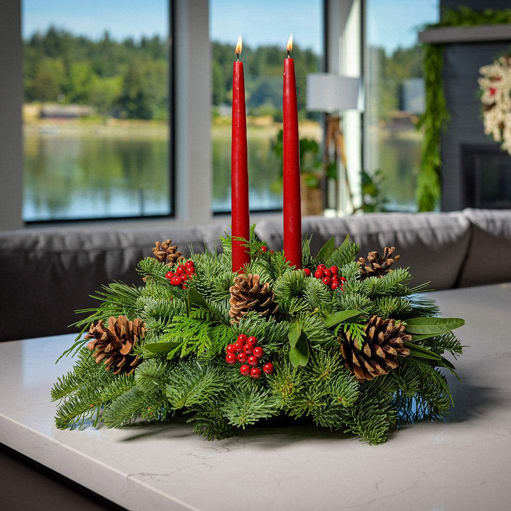 Centerpiece made of noble fir, pine, incense cedar and bay leaves with egg-sized ponderosa pine cones, red berry clusters, and 2 red 12" taper candles on a counter