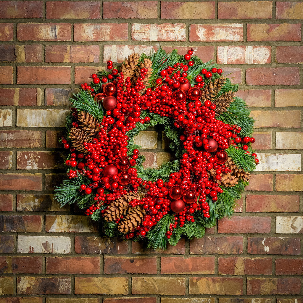 Christmas wreath made of noble fir and white pine with a ring of faux red pepperberries, red ball clusters, and white-pine cones hanging on a brick wall