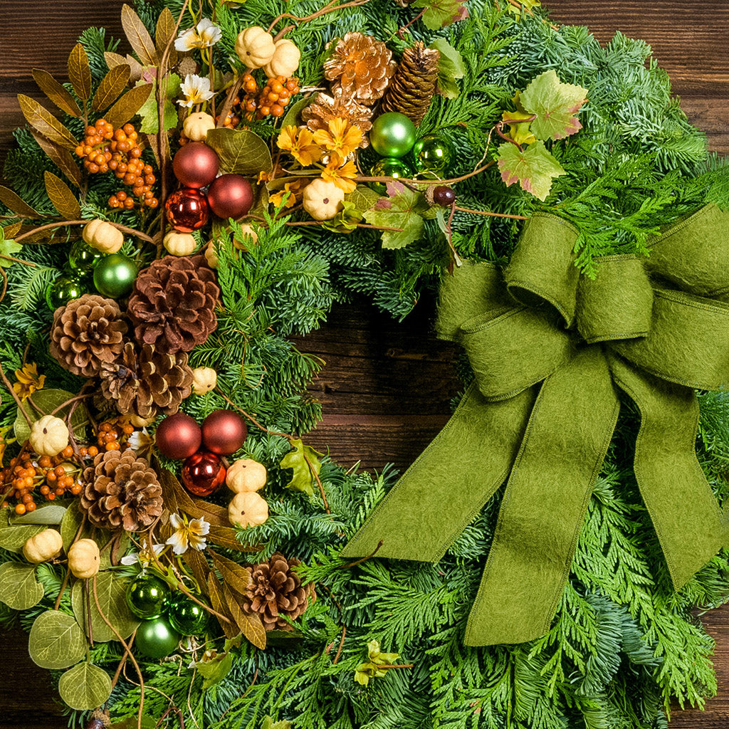 A holiday wreath of noble fir and western red cedar with faux pumpkins, faux acorns, sage accents, leaf and berry accents, 2 copper ball clusters, 2 bright green ball clusters, 3 gold pinecones, 4 Australian pinecones, 1 ponderosa pinecone, and a brushed green linen bow on a wood background.