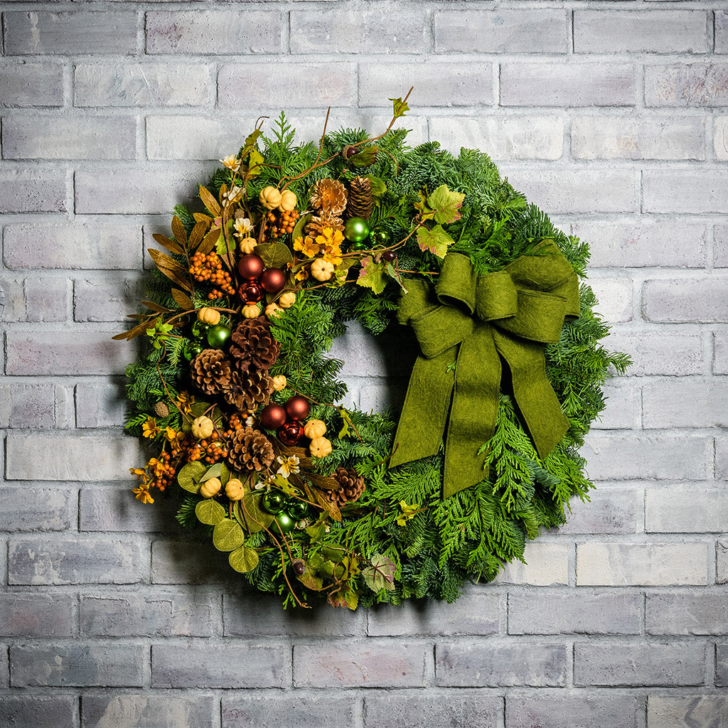 A holiday wreath of noble fir and western red cedar with faux pumpkins, faux acorns, sage accents, leaf and berry accents, 2 copper ball clusters, 2 bright green ball clusters, 3 gold pinecones, 4 Australian pinecones, 1 ponderosa pinecone, and a brushed green linen bow on a white brick background.