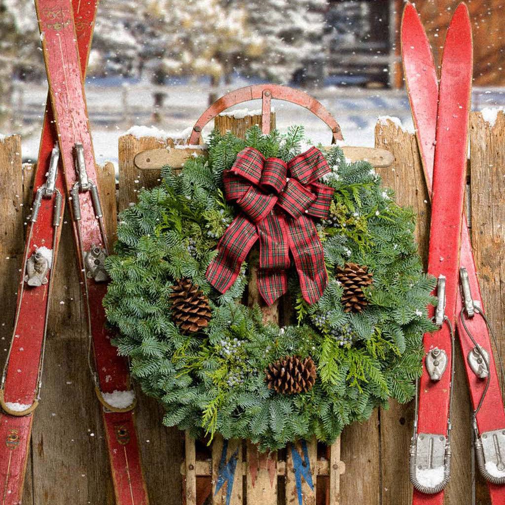 Christmas wreath of noble, cedar, juniper, pine cones and a red plaid bow on a wooden fence near skis and a sled.