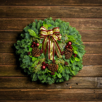 Holiday wreath made of noble fir, incense cedar, juniper, and salal leaves with ponderosa pine cones, faux burgundy berry clusters, ball clusters, and burgundy bow hanging on a wooden wall