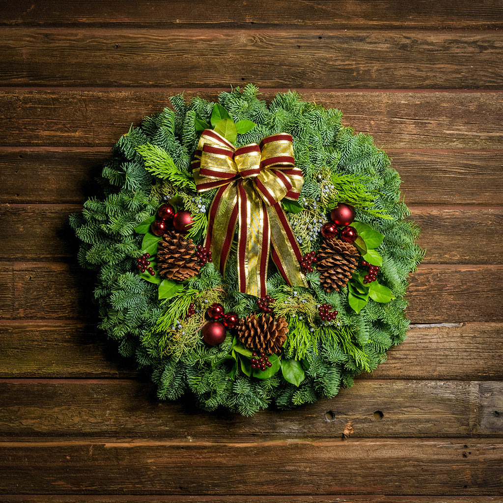Holiday wreath made of noble fir, incense cedar, juniper, and salal leaves with ponderosa pine cones, faux burgundy berry clusters, ball clusters, and burgundy bow hanging on a wooden wall