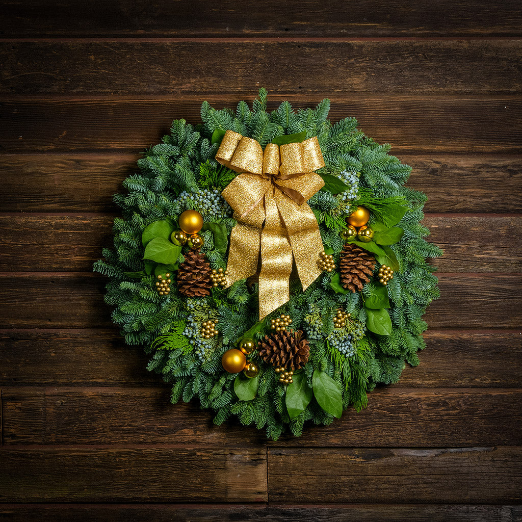 Holiday wreath made of noble fir, incense cedar, juniper, and salal leaves with ponderosa pine cones, faux gold berry clusters, ball clusters, and a gold bow hanging on a wooden wall