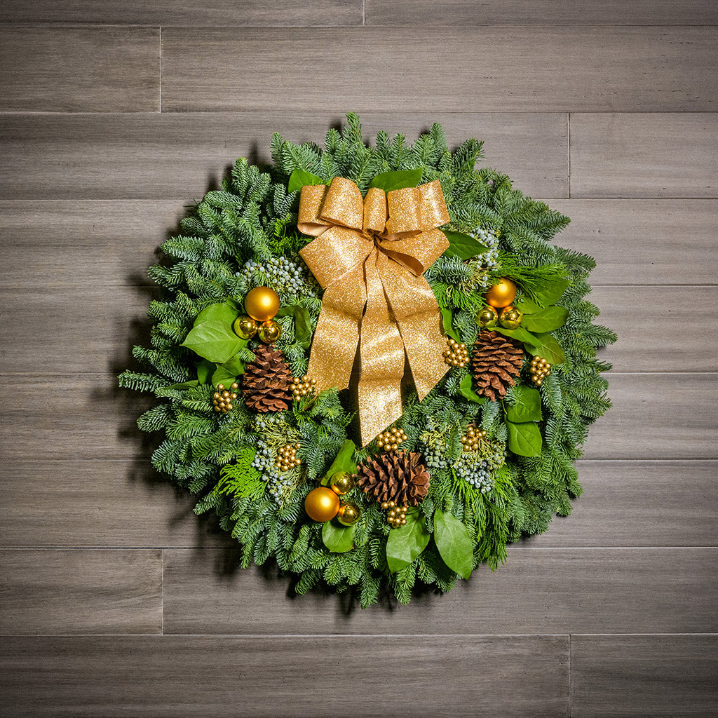 Holiday wreath made of noble fir, incense cedar, juniper, and salal leaves with ponderosa pine cones, faux gold berry clusters, ball clusters, and a gold bow hanging on a wood background