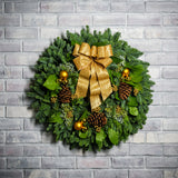 Holiday wreath made of noble fir, incense cedar, juniper, and salal leaves with ponderosa pine cones, faux gold berry clusters, ball clusters, and a gold bow hanging on a brick wall