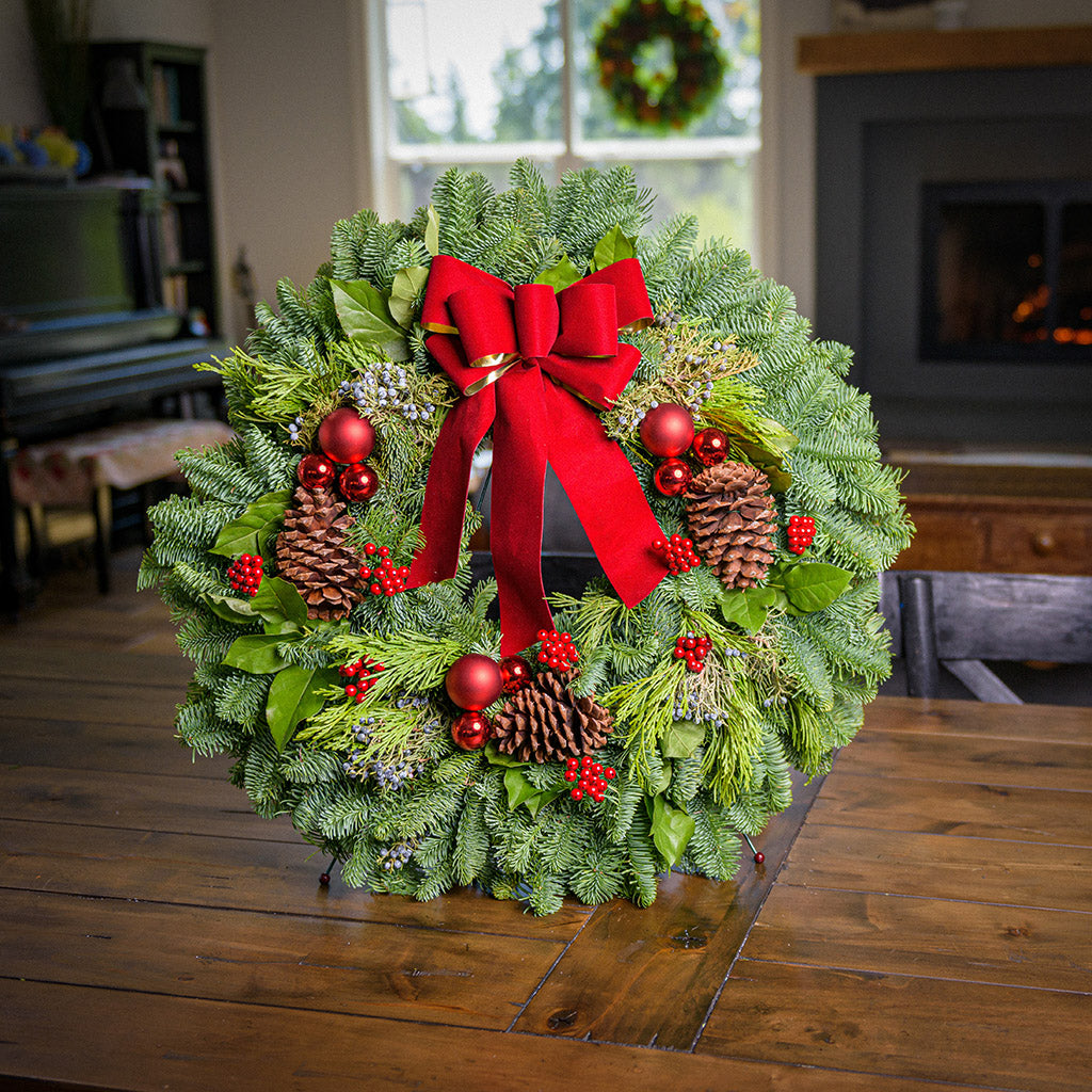 Holiday wreath made of noble fir, incense cedar, juniper, and salal leaves with ponderosa pine cones, faux red berry clusters, ball clusters, and a red bow on wreath stand on a table