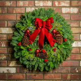 Holiday wreath made of noble fir, incense cedar, juniper, and salal leaves with ponderosa pine cones, faux red berry clusters, ball clusters, and a red bow hanging on a brick wall