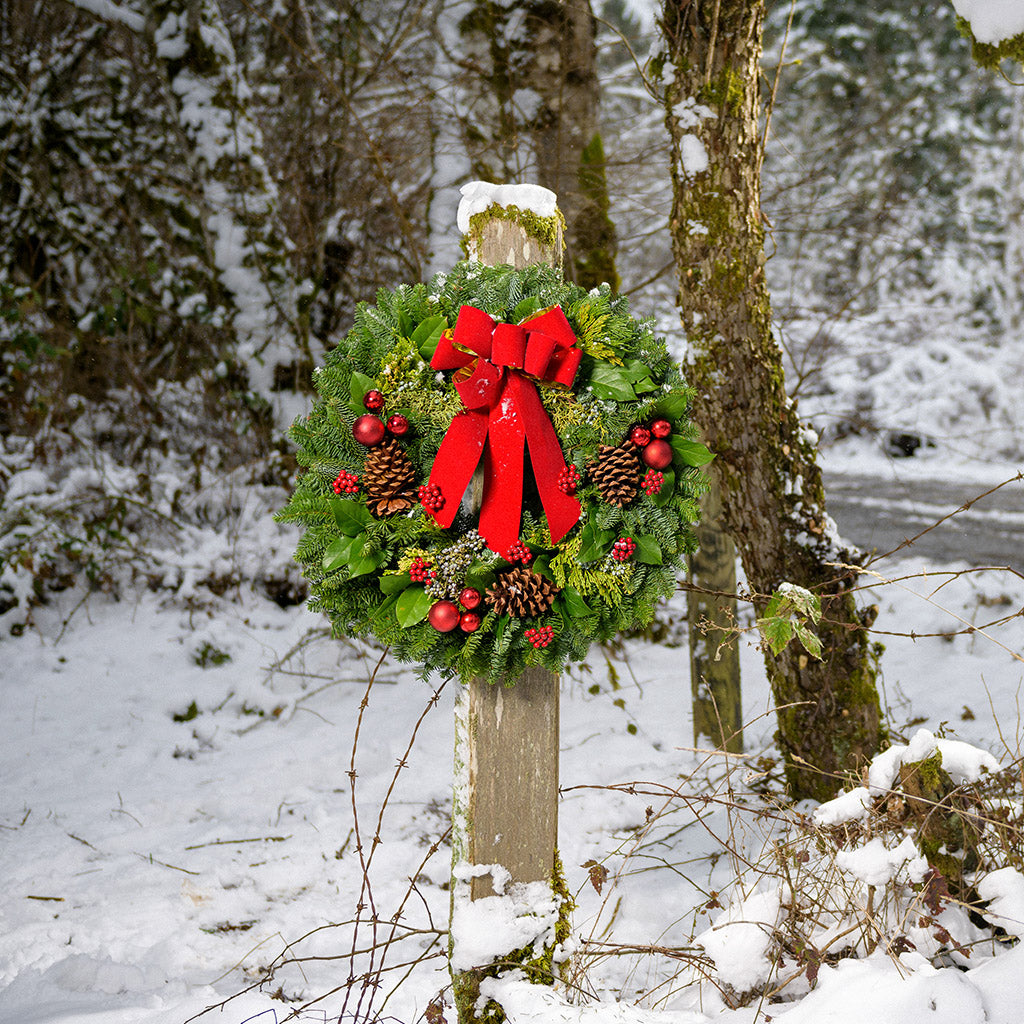 Holiday wreath made of noble fir, incense cedar, juniper, and salal leaves with ponderosa pine cones, faux red berry clusters, ball clusters, and a red bow hanging on a post in a snowy wooded area