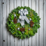 Holiday wreath made of noble fir, incense cedar, juniper, and salal leaves with ponderosa pine cones, faux silver berry clusters, ball clusters, and a silver bow hanging on a white fence