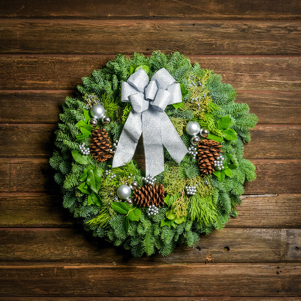 Holiday wreath made of noble fir, incense cedar, juniper, and salal leaves with ponderosa pine cones, faux silver berry clusters, ball clusters, and a silver bow hanging on a wooden wall