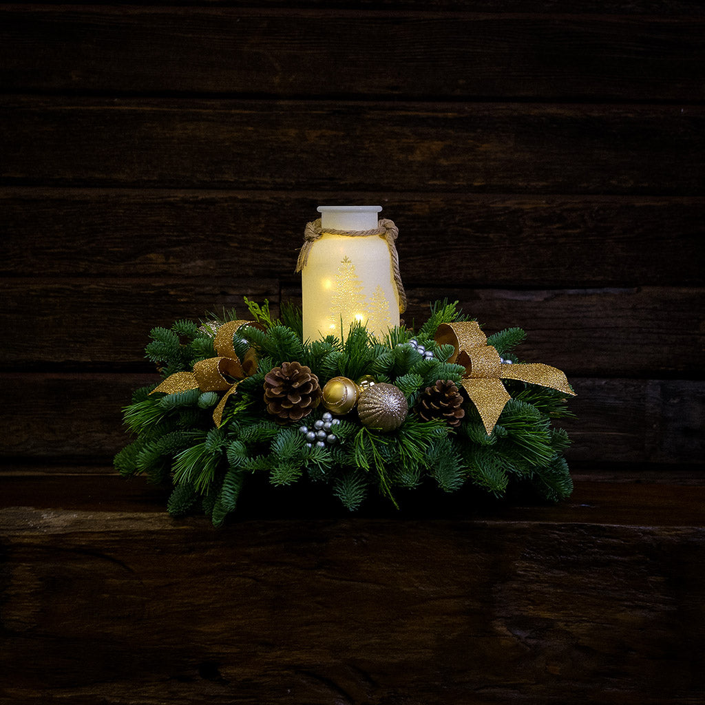 Centerpiece made of noble fir, incense cedar, and white pine with Australian pine cones, gold and silver ball clusters, silver berry clusters, and a lighted holiday glass jar illuminated