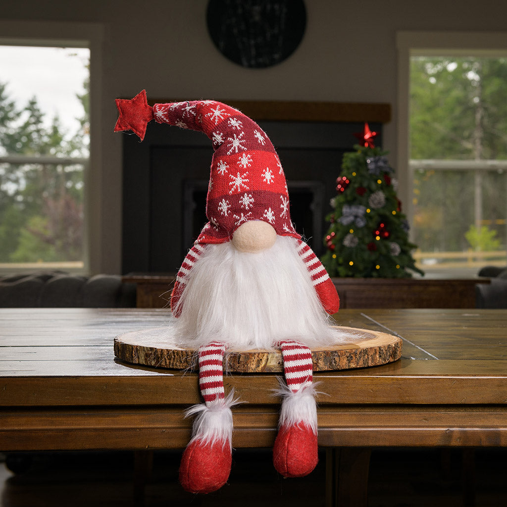 Fluffy white-bearded sitting gnome with red and white knit legs and arms, and nose peeking out from under his pointy bendable red hat covered with white snowflakes and with a star on top sitting on a wood round on a table.