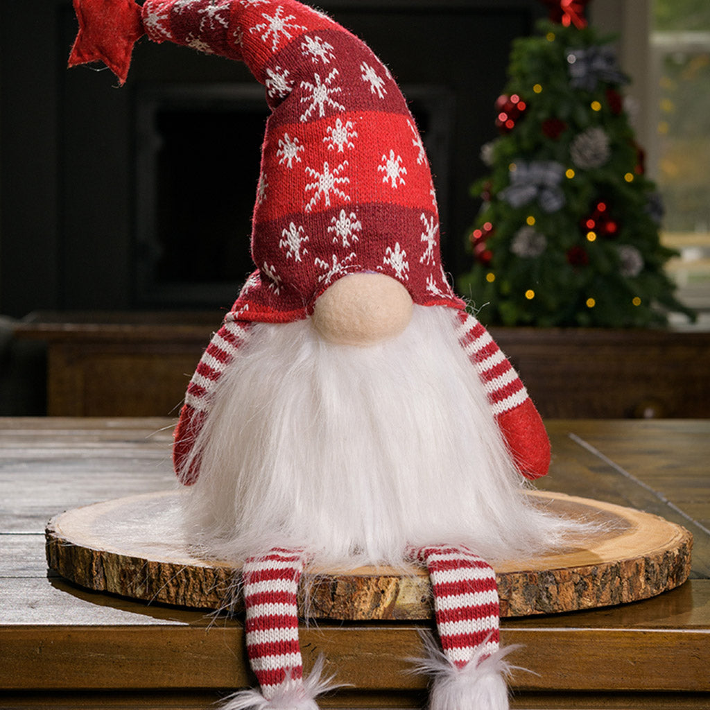 Fluffy white-bearded sitting gnome with red and white knit legs and arms, and nose peeking out from under his  pointy bendable red hat covered with white snowflakes and with a star on top sitting on a wood round on a table. 