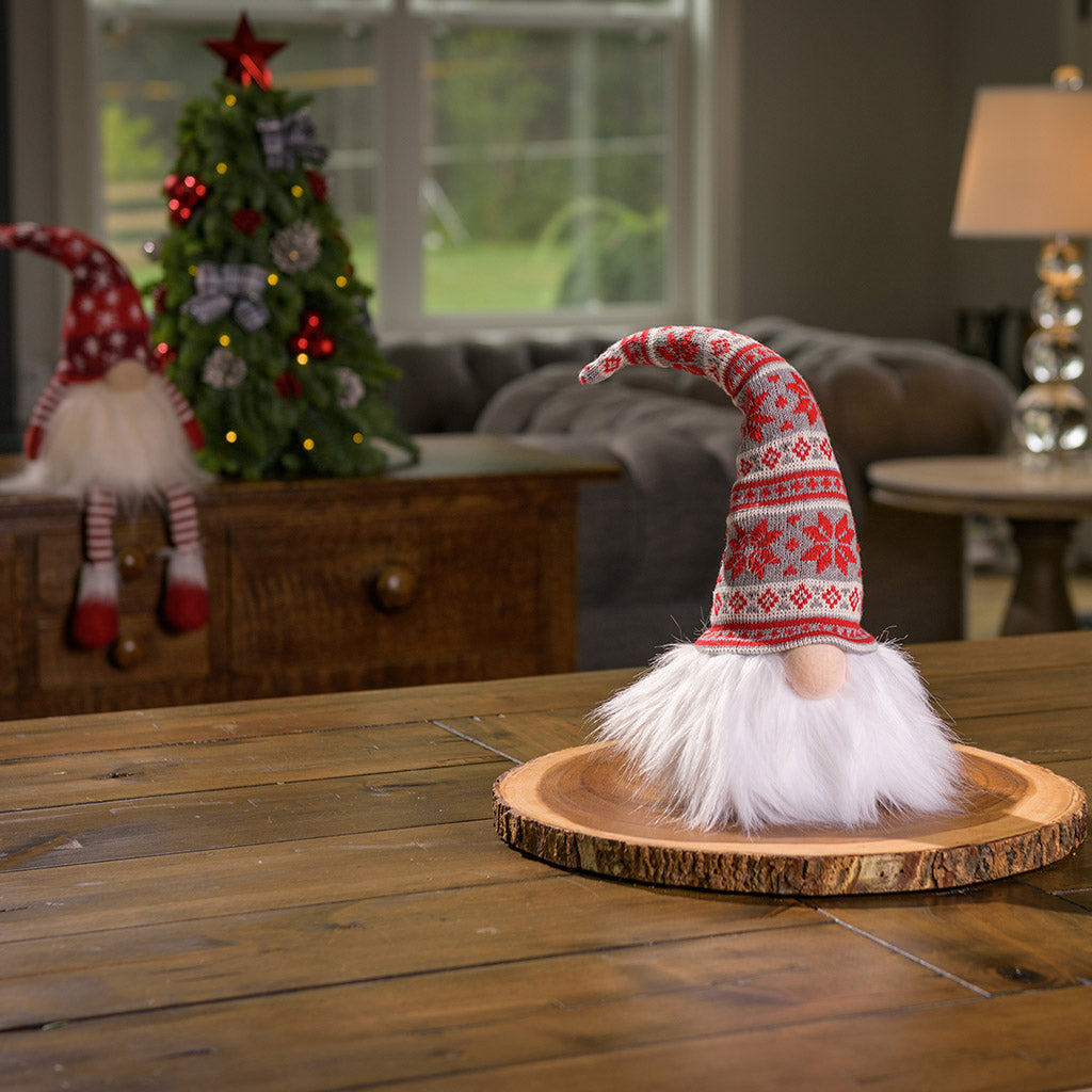 Bearded light up gnome with nose sticking our from under the red, gray, and white Nordic style pointy sweater hat  sitting on a wood round on a table. 