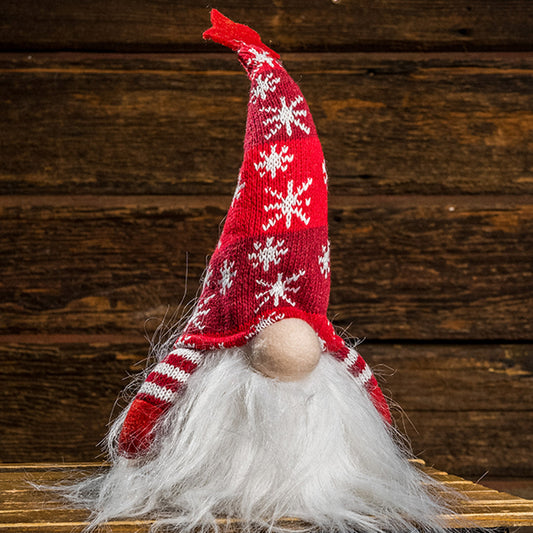 A light up white-bearded gnome with his nose peeking out from under a red knit pointy, bendable hat decorated with snowflakes and a red star on top sitting on a small wood table with a wood background.