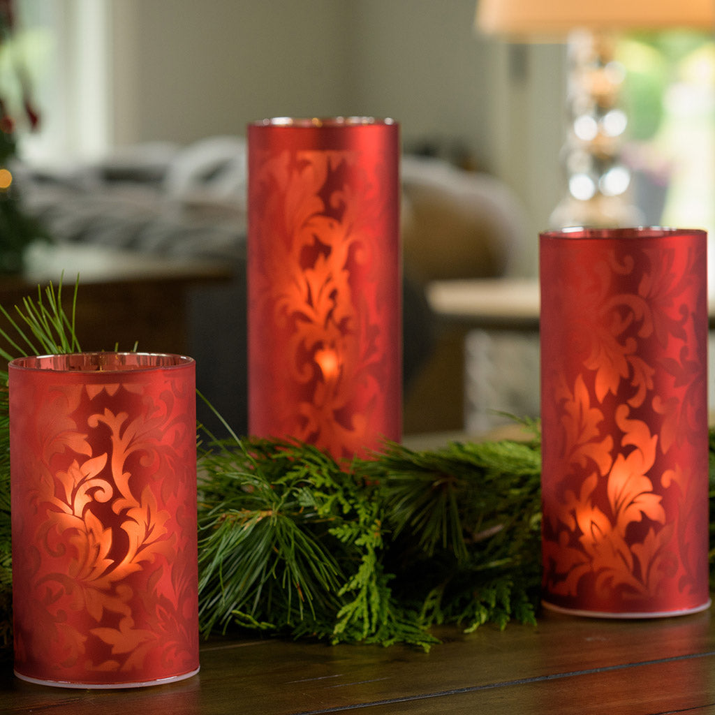 Set of 3 frosted red glass luminaries with a built in timer option paired with garland on a wood table.