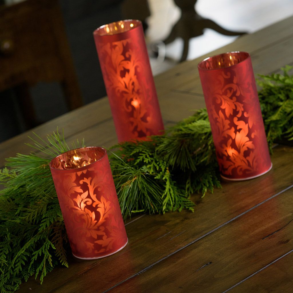 Set of 3 frosted red glass luminaries with a built in timer option paired with garland on a wood table.