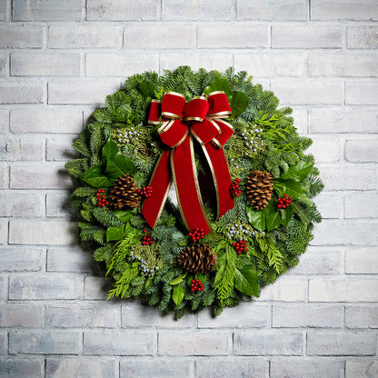 Christmas wreath of noble, cedar, juniper and salal with pine cones, red bow with gold edges and red berry clusters on a white brick background.