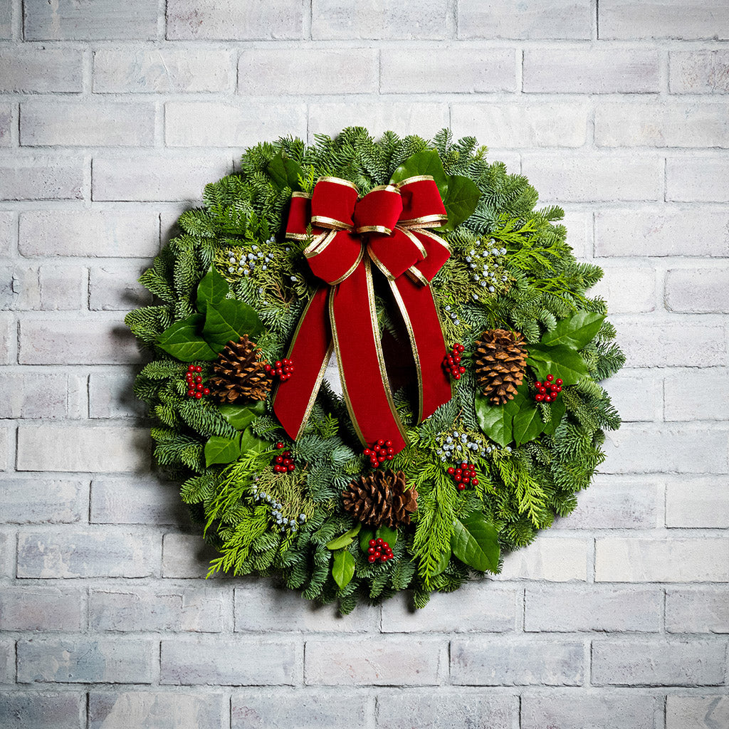 Christmas wreath of noble, cedar, juniper and salal with pine cones, red bow with gold edges and red berry clusters on a white brick background.