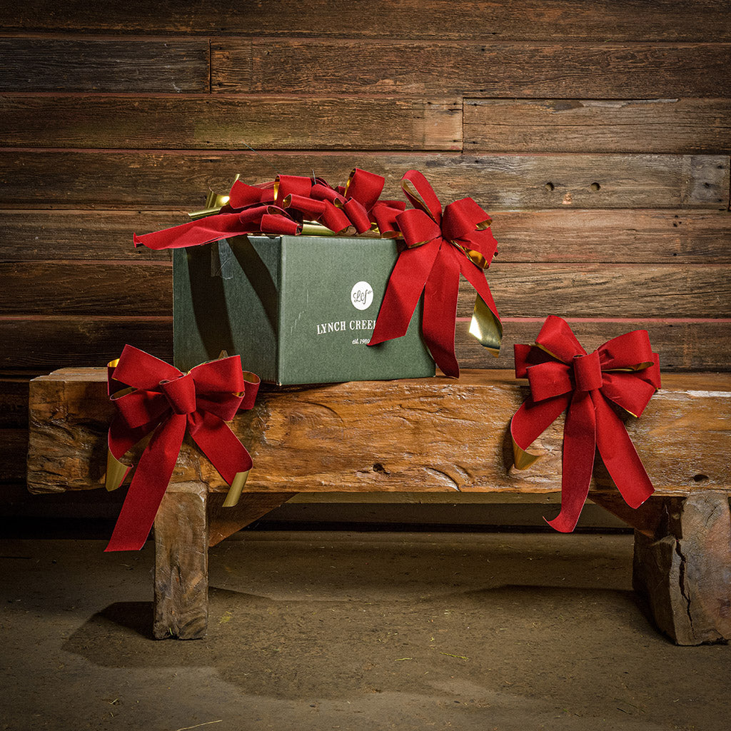 6 Red with shiny gold back bows shown with a green shipping box sitting on a wood bench with a dark wood background.