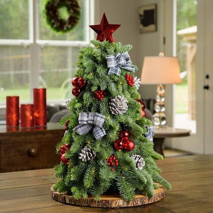 18” miniature tree decorated with white and black plaid bows, shiny red balls and berries, frosted pinecones and topped with a shiny red star tree-topper sitting on a wood round on a wooden table. 