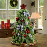 18” miniature tree decorated with white and black plaid bows, shiny red balls and berries, frosted pinecones and topped with a shiny red star tree-topper sitting on a wood round on a wooden table. 