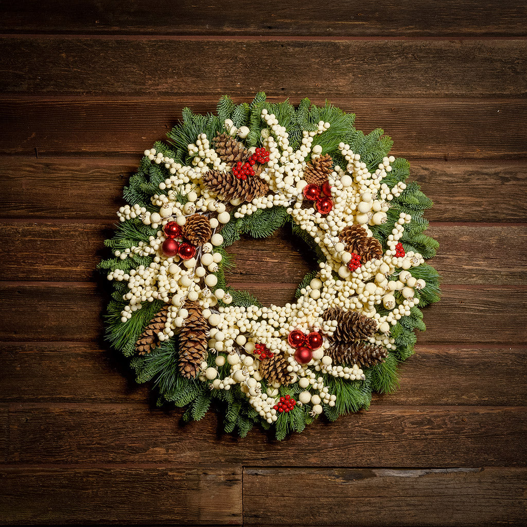 A wreath made of noble fir and pine with cream-colored berries, Australian pinecones, red ball ornaments, and red berry clusters on a dark wooden background.