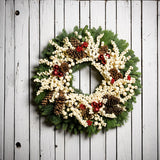 A wreath made of noble fir and pine with cream-colored berries, Australian pinecones, red ball ornaments, and red berry clusters on a white wooden background.