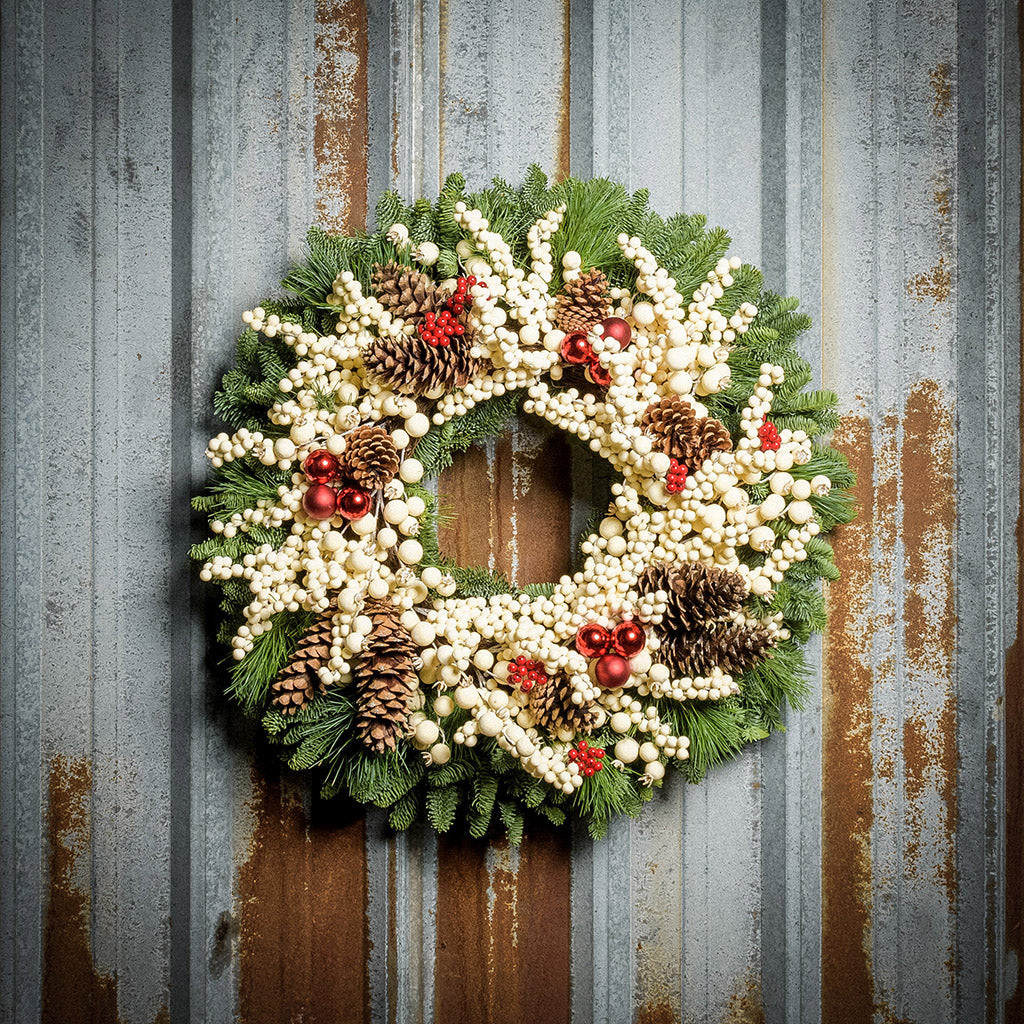 A wreath made of noble fir and pine with cream-colored berries, Australian pinecones, red ball ornaments, and red berry clusters on a rustic metal background.