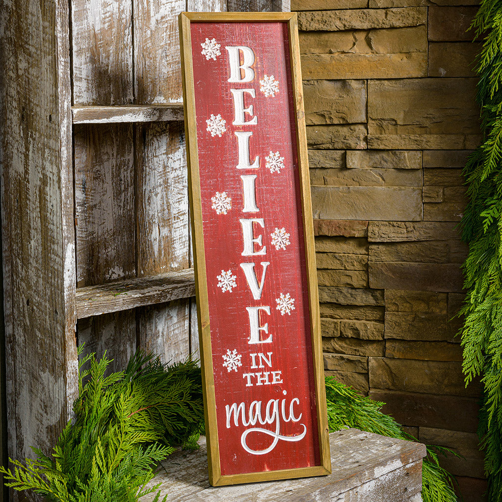 Framed wood sign engraved with "Believe in the Magic" in white while falling snowflakes surround the words on a front porch.