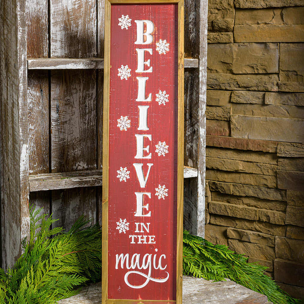 Framed wood sign engraved with "Believe in the Magic" in white while falling snowflakes surround the words on a front porch.