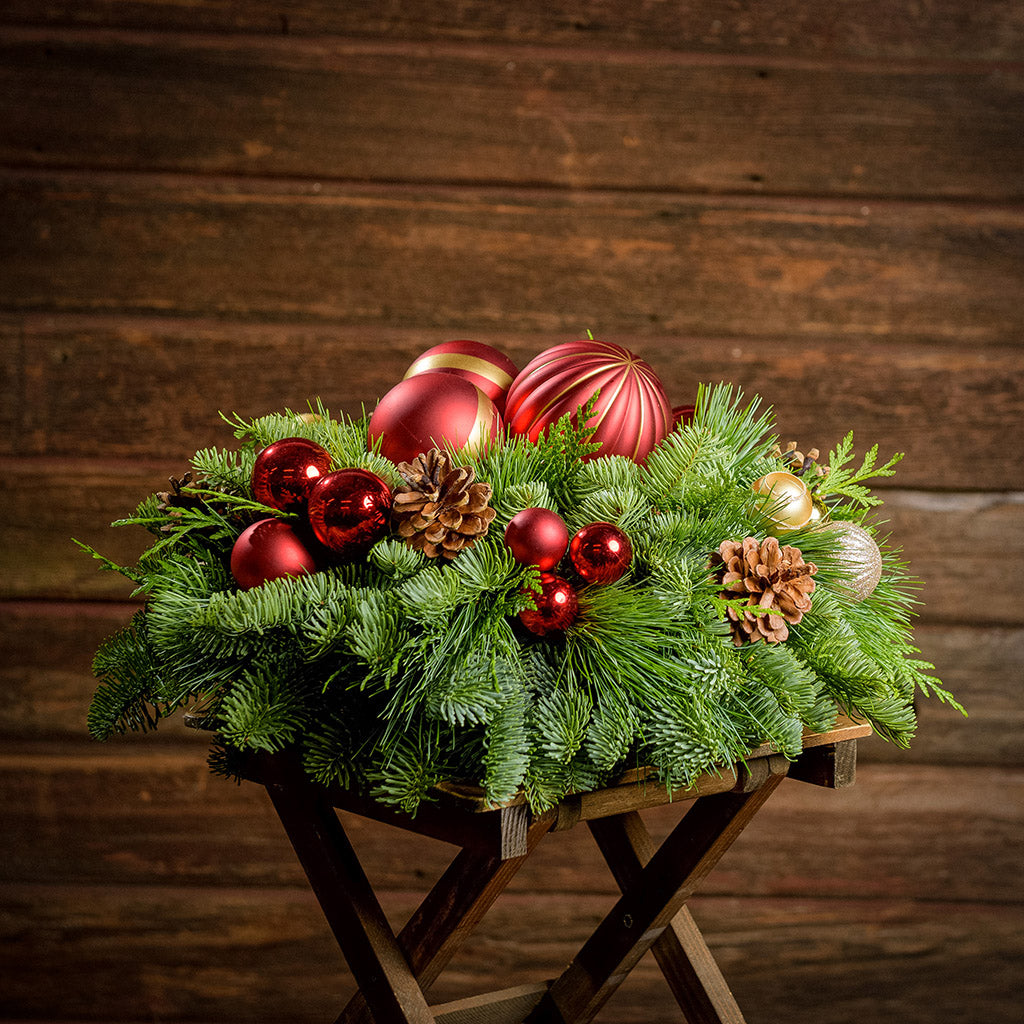 An arrangement made of noble fir, incense cedar, and white pine with Austrian pinecones, red and gold ball clusters, and large red ball ornaments sitting on a wood table with a dark wooden background.