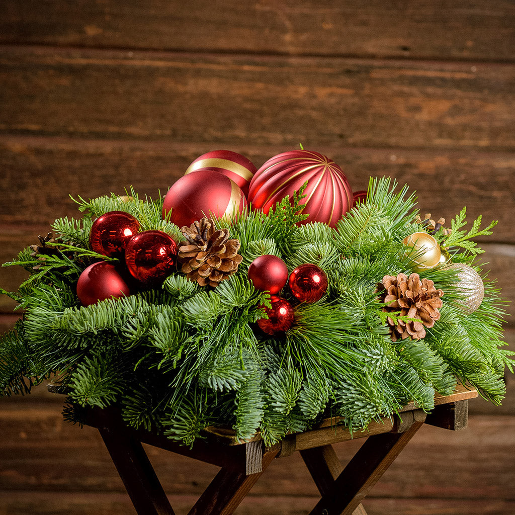 An arrangement made of noble fir, incense cedar, and white pine with Austrian pinecones, red and gold ball clusters, and large red ball ornaments sitting on a wood table with a dark wooden background.
