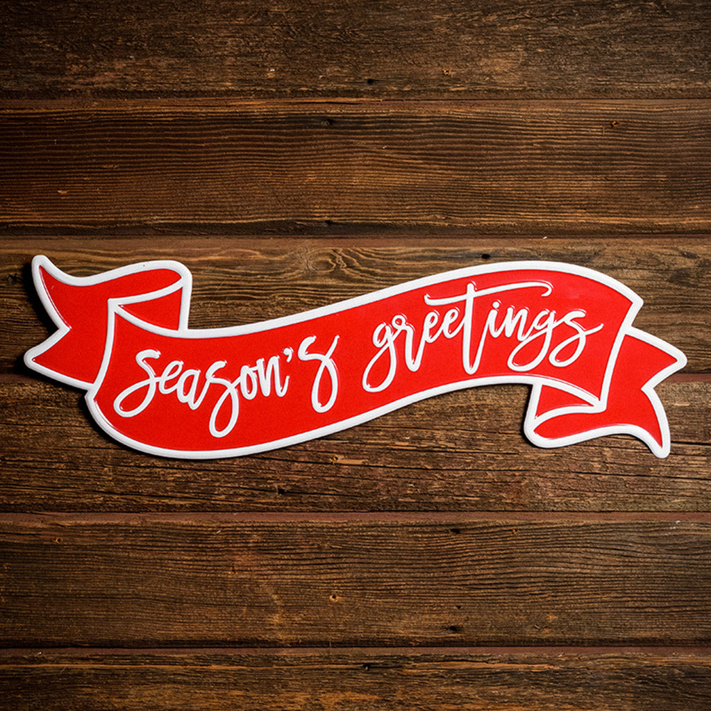 24" red and white metal Season's greetings sign on a dark wooden background.