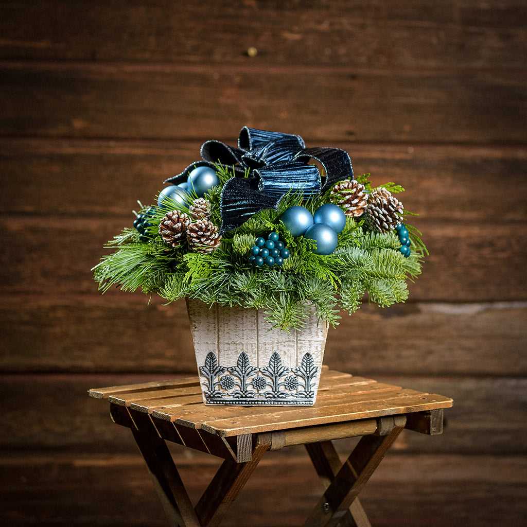 An arrangement made of noble fir, incense cedar, and white pine with dark blue berries, mini frosted pinecones, dark blue ball clusters, and a midnight blue bow in a whitewashed wood container with a dark wood background.
